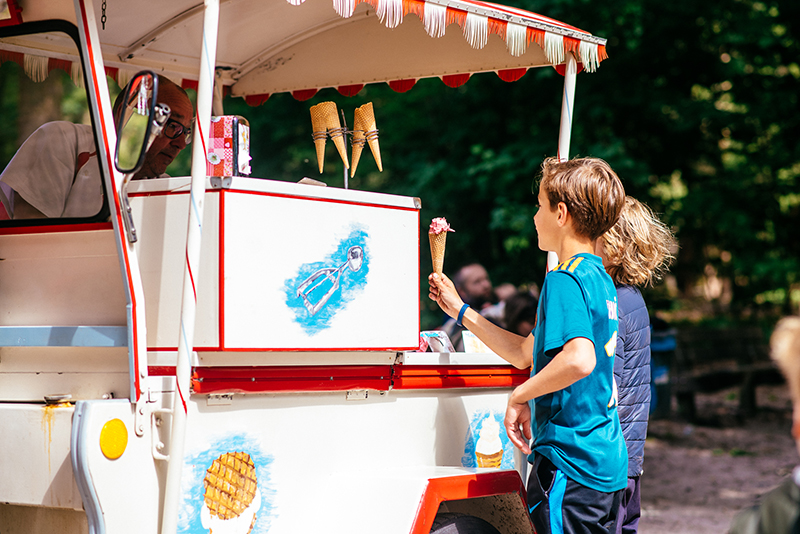 Twee kinderen staan naast elkaar bij een leuke witte ijskar en een van deze kinderen krijgt een ijsje aangeboden van de ijscoman.